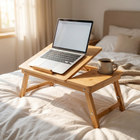 Bamboo laptop bed table set up on a bed with an open laptop on the tilted panel and a coffee mug in the cup holder.