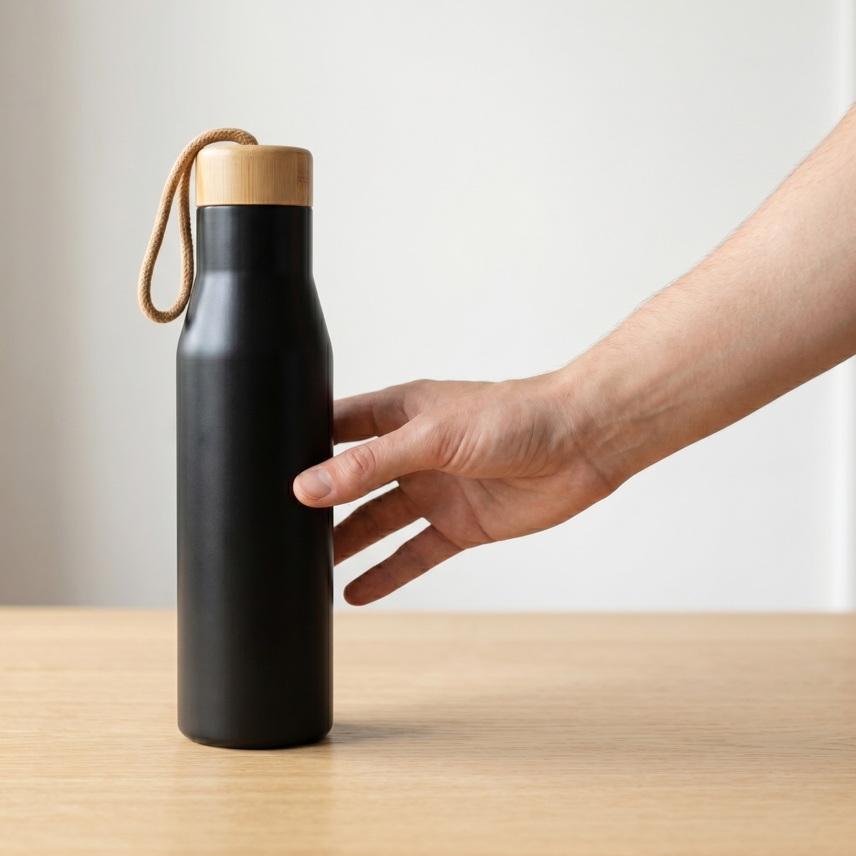 Hand reaching for the Bamboo Steel Travel Bottle on a pale oak desk — editorial minimal product moment