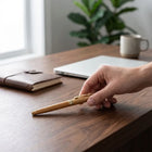 Hand placing a Bamboo Roller Pen on a wooden desk with a notebook and laptop in the background.