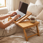 Person typing on a laptop on the tilted bamboo desk in bed, tea mug in the cup holder, showing the adjustable tilt.